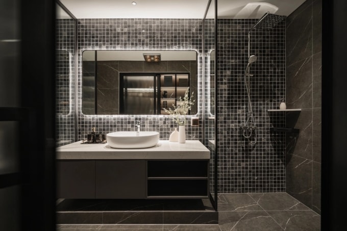Phòng tắm sử dụng tông màu tối làm chủ đạo, kết hợp gạch mosaic và đá vân tự nhiên, tạo cảm giác sang trọng, sâu lắng nhưng không gây bí bách nhờ hệ đèn hắt quanh gương và bố cục chiếu sáng hợp lý. Ảnh: KLUX