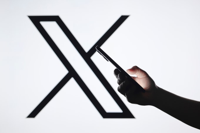 Một người dùng smartphone, phía sau là logo mạng xã hội X. Ảnh: Reuters