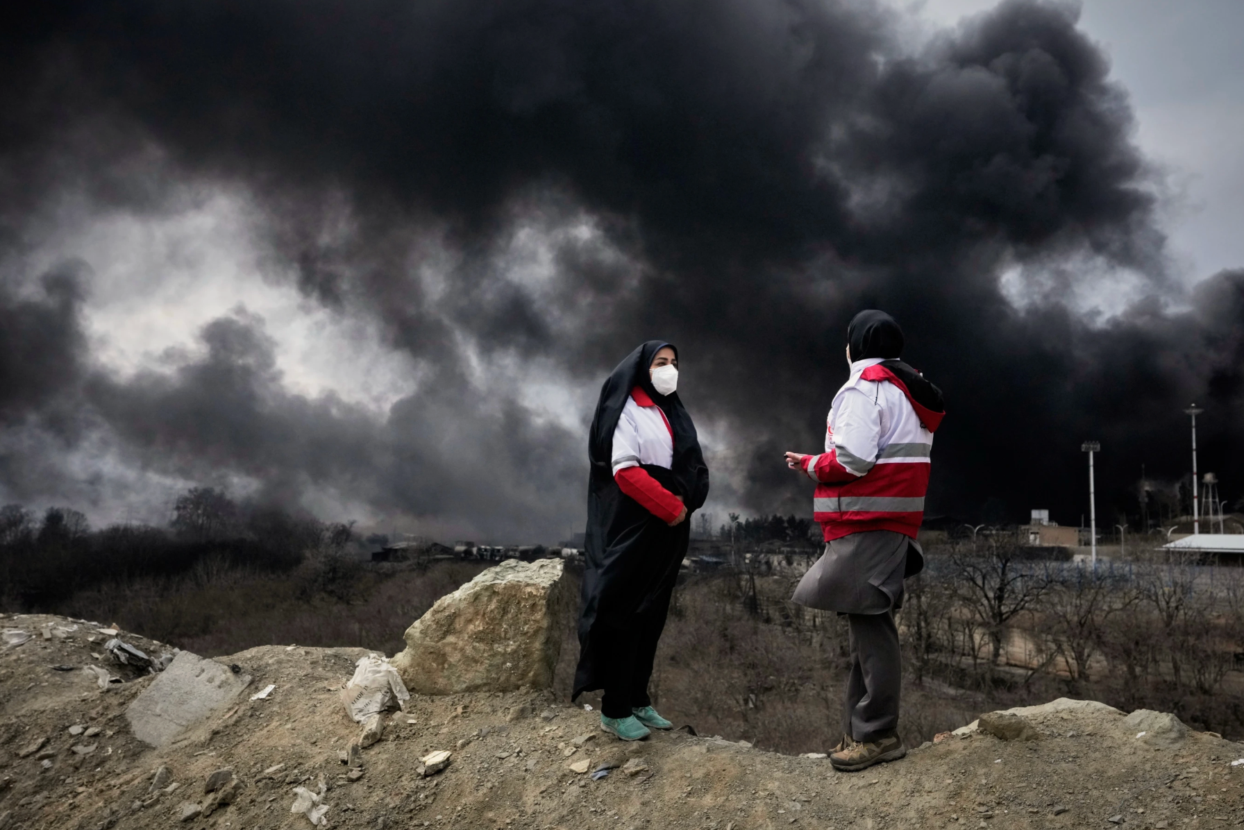 Hai người phụ nữ thuộc Hội Trăng lưỡi đỏ Iran đứng khi cột khói dày đặc bốc lên từ vụ tấn công của Mỹ và Israel vào một kho chứa dầu ở Tehran, Iran. (Ảnh AP/Vahid Salemi)